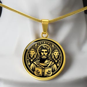 May include: Gold-tone pendant necklace featuring a round pendant with a black and gold design. The pendant depicts a figure with a halo, holding a book, flanked by two lions. The necklace has a delicate chain.