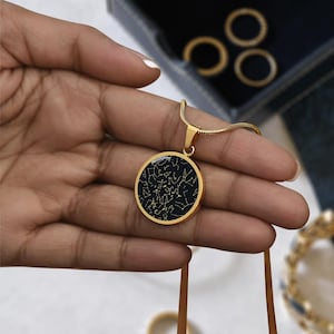 May include: A gold-colored pendant necklace featuring a round pendant with a black background and a constellation design in gold. The necklace is held in a hand, with a gold chain and clasp visible. The pendant is approximately 1.25 inches in diameter.