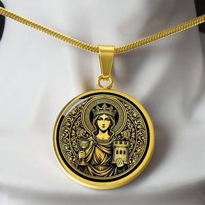May include: A gold-toned circular pendant necklace featuring a black and gold graphic illustration of a crowned figure holding a chalice and a castle tower. The pendant hangs from a matching gold-toned snake chain, showcasing intricate filigree details.
