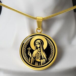 May include: A gold-colored pendant necklace featuring a round pendant with a religious icon. The icon depicts a woman holding a cross and a book, set against a black background with gold accents. The necklace chain is also gold.