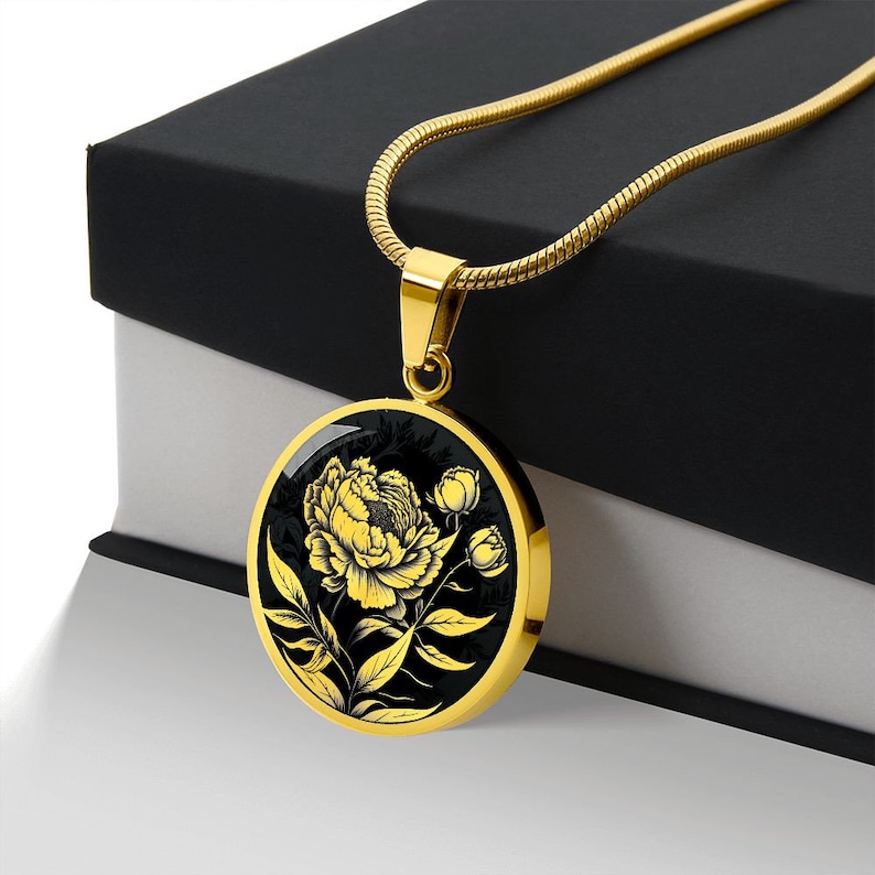 May include: A gold chain necklace with a round pendant featuring a black and gold floral design. The pendant depicts a single peony flower with leaves and buds.