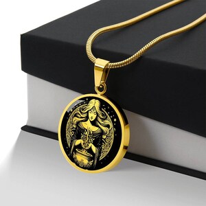 May include: A gold necklace with a round pendant featuring a black and gold illustration of a woman holding a cauldron and herbs. The woman has long flowing hair and is surrounded by a crescent moon and stars.