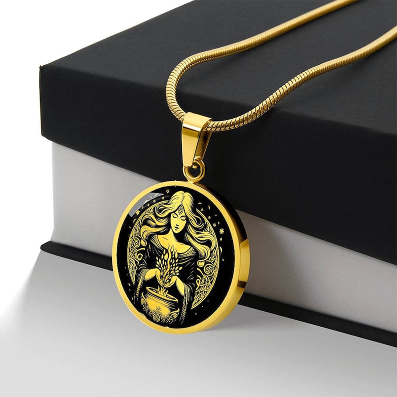 May include: A gold necklace with a round pendant featuring a black and gold illustration of a woman holding a cauldron and herbs. The woman has long flowing hair and is surrounded by a crescent moon and stars.