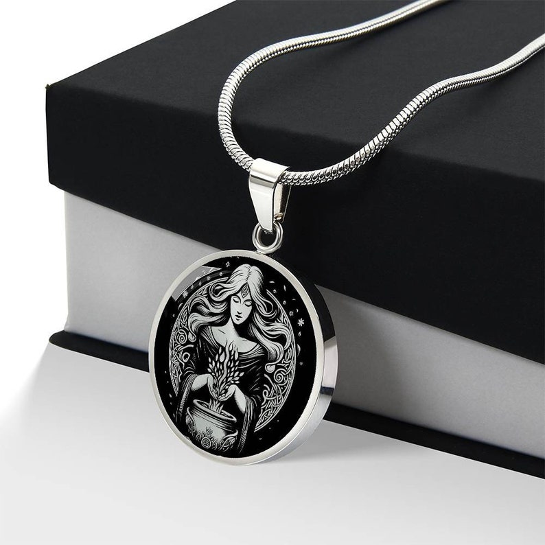 May include: A silver necklace with a round pendant featuring a black and white illustration of a woman holding a cauldron with a plant growing from it. The woman is surrounded by a circle of vines and stars.