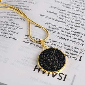 May include: A gold-colored necklace with a circular pendant featuring a black background and a constellation design in gold. The pendant is on a thin, gold chain, and the necklace is displayed on a book.