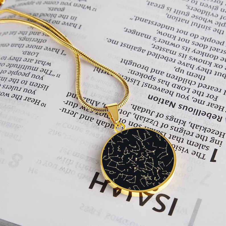 May include: A gold-colored necklace with a circular pendant featuring a black background and a constellation design in gold. The pendant is on a thin, gold chain, and the necklace is displayed on a book.