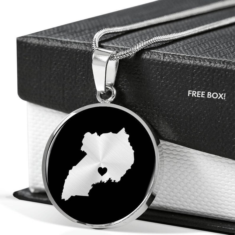May include: A silver necklace with a round pendant featuring a black and white outline of Uganda with a small heart in the center. The necklace is displayed in a black box with the words "FREE BOX!" printed on it.
