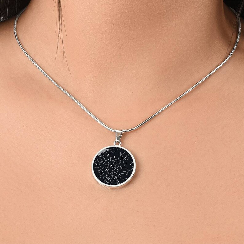 May include: A silver-toned necklace featuring a round pendant with a dark background and a white constellation design. The pendant is suspended from a delicate chain, creating a celestial-themed accessory.