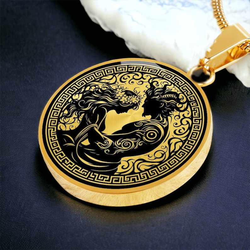 May include: A gold pendant necklace with a circular design featuring a black and white silhouette of two figures embracing. The pendant is surrounded by a decorative border of a repeating Greek key pattern.