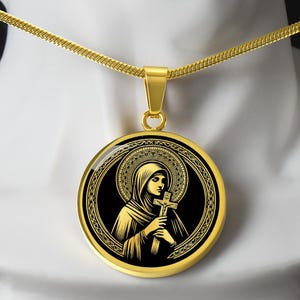 May include: Gold-toned pendant necklace featuring a religious icon. The round pendant showcases a detailed illustration of a figure holding a cross, set against a black background with gold accents. The necklace has a delicate chain.