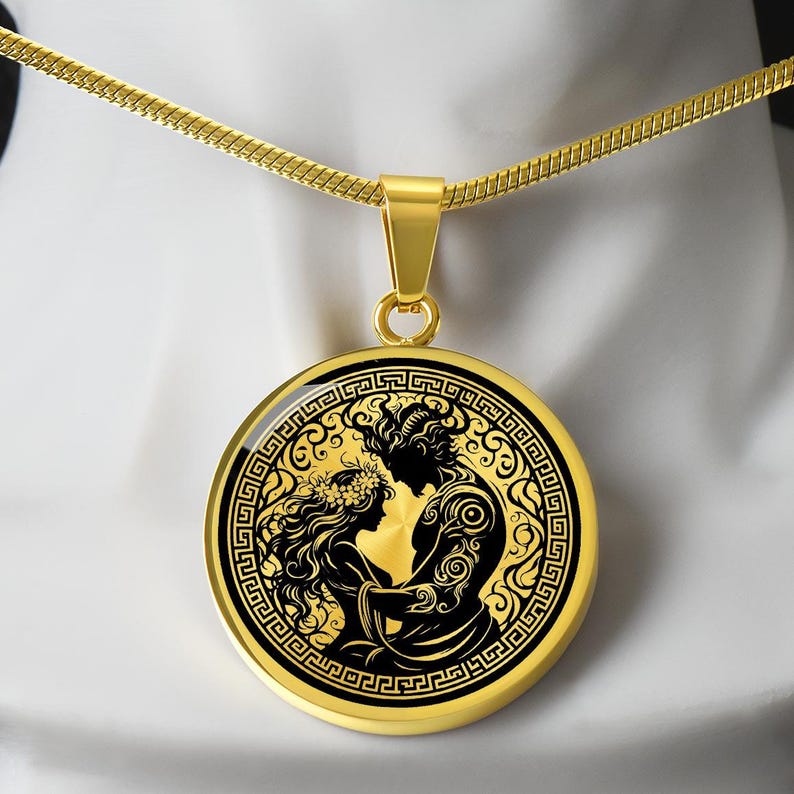 May include: A gold chain necklace with a round pendant featuring a black and gold design of a couple embracing. The pendant has a Greek key border.