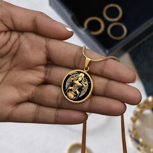 May include: Gold necklace with a round pendant featuring a black and gold illustration of a woman holding a cauldron. The pendant is surrounded by a gold border.
