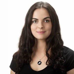 May include: A woman with long dark brown hair wearing a black t-shirt and a silver necklace with a black pendant. The pendant is a round disc with a white design.