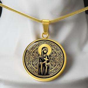 Può includere: Una collana con pendente dorato raffigurante un'icona religiosa. Il pendente rotondo presenta un disegno nero e oro di una figura che tiene una croce, circondata da un bordo ornato. La catena della collana è una sottile catena a serpente dorata.