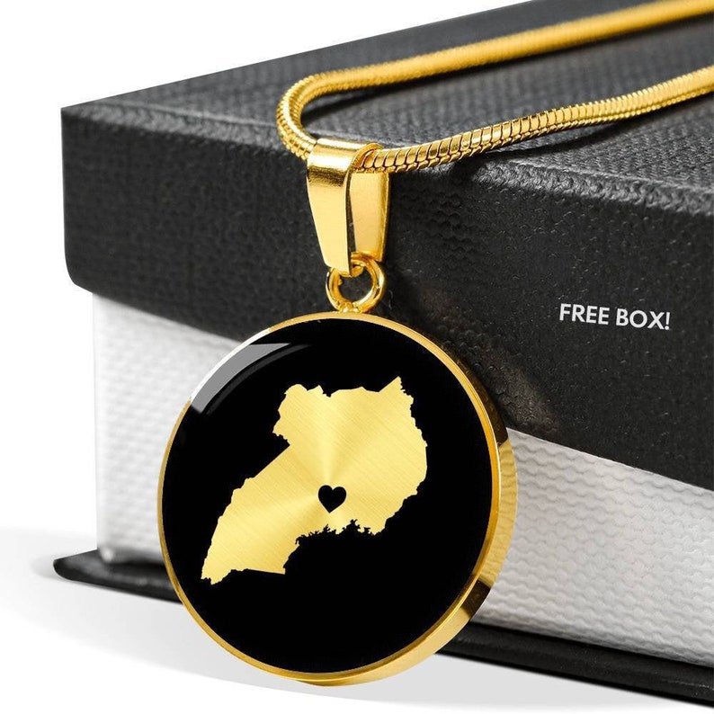 May include: Gold necklace with a round pendant featuring a gold outline of Uganda with a small heart in the center. The pendant is on a black background. The necklace is in a black box with the words "FREE BOX!" printed on it.