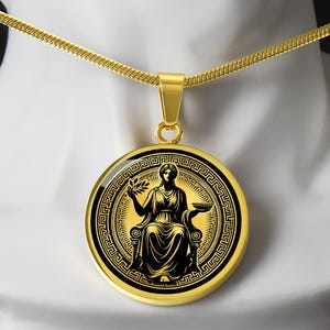 May include: A gold necklace with a round pendant featuring a black silhouette of a woman holding a branch with leaves and a bowl. The pendant is surrounded by a black and gold Greek key pattern.