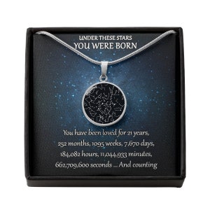 May include: A silver necklace with a round pendant featuring a star map design. The pendant hangs on a thin chain, presented in a black box with text that reads "UNDER THESE STARS YOU WERE BORN".