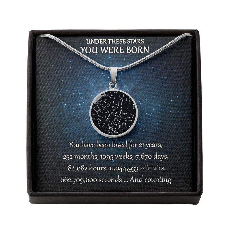 May include: A silver necklace with a round pendant featuring a star map design. The pendant hangs on a thin chain, presented in a black box with text that reads "UNDER THESE STARS YOU WERE BORN".