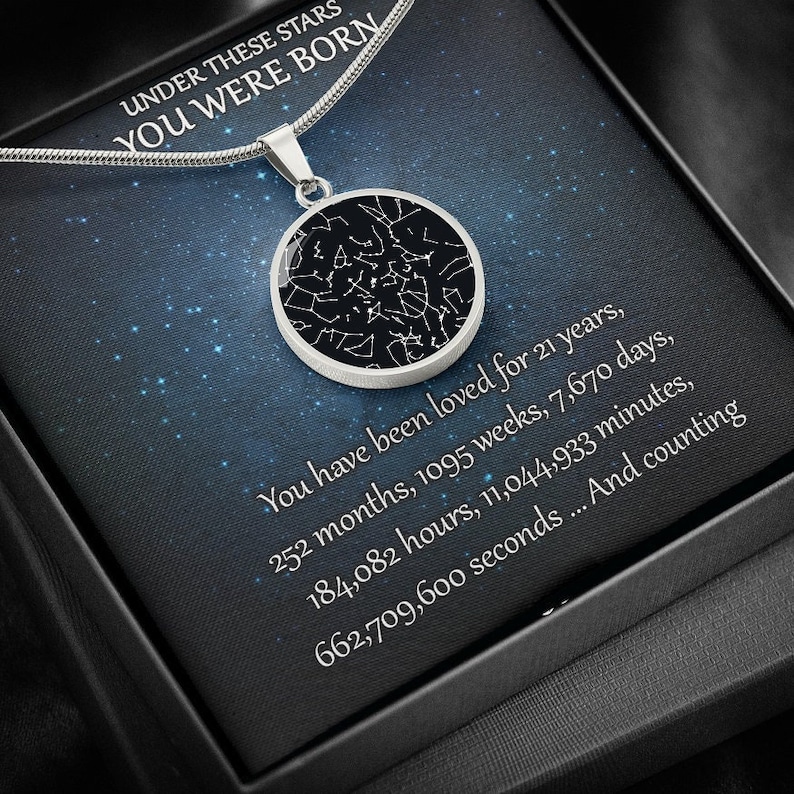 May include: A silver-colored pendant necklace with a round charm featuring a black background and a white constellation design. The necklace is presented in a black box with text that reads "UNDER THESE STARS YOU WERE BORN."