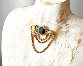 May include: A vintage gold-tone brooch with a dark green oval center stone and three dangling chains. The brooch has ornate detailing and is displayed on a white mannequin bust.
