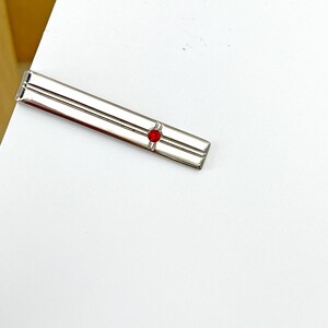 May include: A silver-toned metal tie bar featuring a long, rectangular design with two parallel horizontal grooves running along its length and a single, small, circular red gemstone set prominently near one end of the accessory.