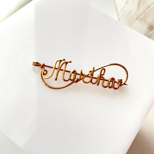May include: A gold-coloured wire name pin spelling "Martha" in cursive. The pin is a delicate piece of jewellery, with a pin clasp on the back. The pin is on a white surface.