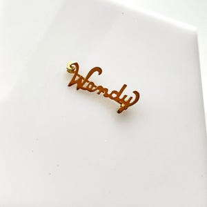 Puede incluir: Un broche de metal dorado con la forma del nombre "Wendy" en fuente cursiva. El broche está sobre un fondo blanco. El broche es pequeño y delicado, con un acabado brillante.