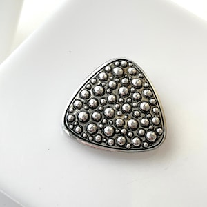 Puede incluir: Broche triangular plateado con una superficie texturizada de pequeños detalles circulares en relieve. El broche tiene un aspecto metálico pulido y está sobre un fondo blanco.