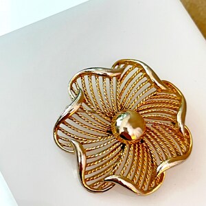 May include: A gold-tone metal brooch shaped like a five-petaled flower. Each petal features a textured, twisted wire design radiating from a smooth, polished, circular center. The edges of the petals are finished with a solid, smooth gold-tone border.