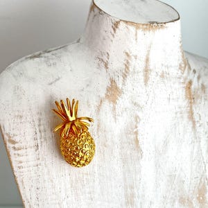 Könnte beinhalten: Eine goldfarbene Ananas-Brosche. Die Ananas ist mit strukturierten Blättern und einem strukturierten Körper verziert.
