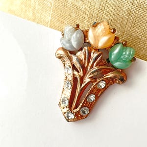 May include: Vintage gold-tone brooch featuring a floral design with three flower-shaped accents in gray, peach, and green. The brooch has clear rhinestone accents and a decorative leaf-shaped base.