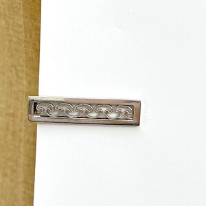 May include: A rectangular silver-toned tie bar featuring a cut-out design of repeating wave patterns. The metal accessory has a polished finish and a clean, linear frame that encloses the stylized wave motif, creating a balanced and symmetrical appearance.