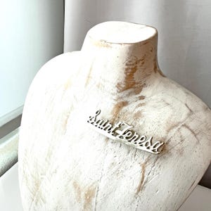 May include: A silver-colored brooch with the name "Saint Teresa" in a cursive font. The brooch is displayed on a white mannequin bust. The brooch is a decorative accessory.