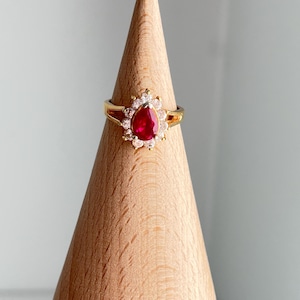 May include: A gold ring featuring a pear-shaped, deep red gemstone surrounded by smaller, clear stones. The ring is set on a wooden display, showcasing its intricate design and vibrant color.