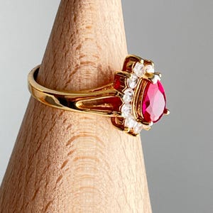 Puede incluir: Un anillo dorado con una piedra preciosa roja en forma de pera, rodeada de piedras claras más pequeñas. La banda del anillo es dorada pulida y el engaste tiene un diseño ornamental en forma de flor. El anillo se exhibe sobre un soporte de madera.