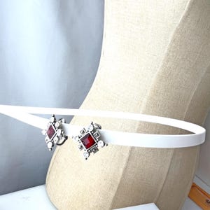 May include: A white belt with two silver-toned diamond-shaped embellishments. Each embellishment features a central red gemstone surrounded by smaller clear stones. The belt is displayed on a mannequin.