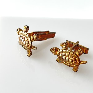 May include: A pair of gold-toned cufflinks featuring turtle designs. The cufflinks have a detailed turtle shape with textured shells and small legs. The metal has a slightly aged appearance, adding to their vintage charm.