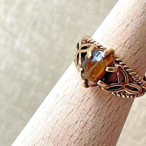 May include: A gold-toned ring featuring a central oval-shaped brown and amber tiger eye gemstone held by four prongs. The band displays an intricate openwork design with twisted rope-like edges and symmetrical cutouts on both sides of the stone.