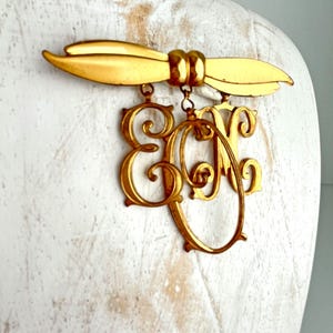 May include: A gold-toned brooch featuring a stylized leaf design at the top, with the letters "E" and "M" in a decorative script hanging below. The brooch is displayed on a white surface.
