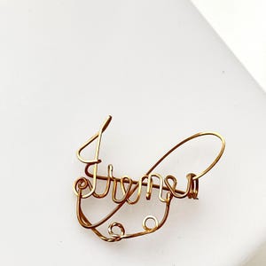 Puede incluir: Una pieza de arte de alambre de color dorado que deletrea la palabra "Dreams" en escritura cursiva. El alambre está doblado y moldeado para formar las letras, creando un diseño delicado y artístico. La pieza está sobre un fondo blanco.