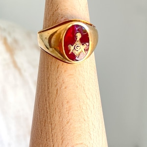 Puede incluir: Un anillo dorado con una cara rectangular con un centro rojo. El centro muestra un símbolo masónico en oro. El anillo se exhibe sobre un soporte de madera, mostrando su diseño y artesanía.