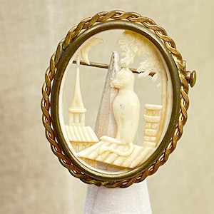 Puede incluir: Un broche ovalado con un marco de metal dorado trenzado que rodea una escena tallada de color crema. El relieve muestra a un gato sentado en un tejado junto a una chimenea y la torre de una iglesia, con una luna creciente y un árbol de fondo.