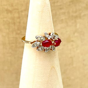 May include: A gold-toned ring featuring two prominent oval-cut red stones arranged horizontally. The setting is accented with small, clear, round-cut stones arranged in a leaf-like pattern surrounding the central red gems on a thin metal band.