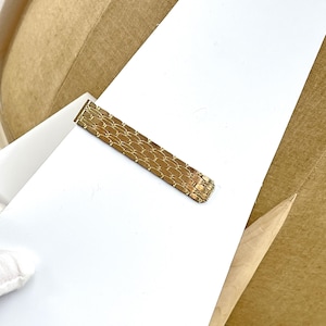 May include: A gold-toned metal tie bar featuring a textured, woven-style link pattern across its rectangular surface. The accessory has a polished finish and a straight, elongated design with rounded edges at one end, showcasing a repeating scale-like motif.