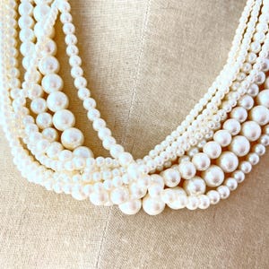 Puede incluir: Collar de perlas de múltiples hilos con cuentas redondas de color blanco roto de varios tamaños. Los hilos se superponen, creando un efecto de capas. Las perlas tienen un brillo sutil, sobre un fondo de tela neutra.