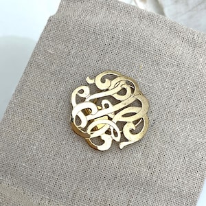 May include: A circular gold-toned brooch featuring an intricate, openwork design of swirling, interconnected filigree patterns. The metallic surface has a polished finish, creating a reflective appearance across the curved, decorative metal lines.