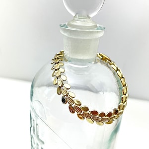 May include: A gold-toned bracelet with a leaf-shaped chain design. The bracelet is displayed on a clear glass bottle.