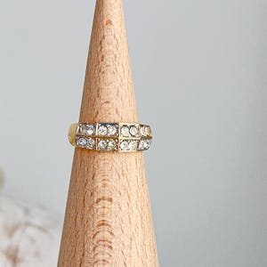 May include: A gold-toned ring with two rows of clear, sparkling stones. The ring is displayed on a wooden cone-shaped stand against a soft gray background. The ring's design features a classic, elegant style.
