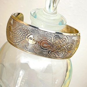 May include: A silver-toned metal cuff bracelet featuring an embossed design of a fish swimming among stylized waves and a central intricate knot pattern. The bracelet has a polished finish and an open back, showcasing detailed engravings across the surface.