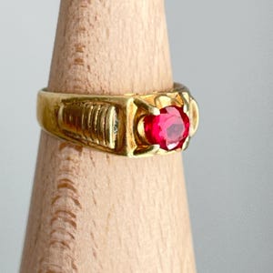 May include: A gold ring with a large, round, red gemstone. The ring has a rectangular face with a ribbed design on one side. The ring is displayed on a wooden stand.